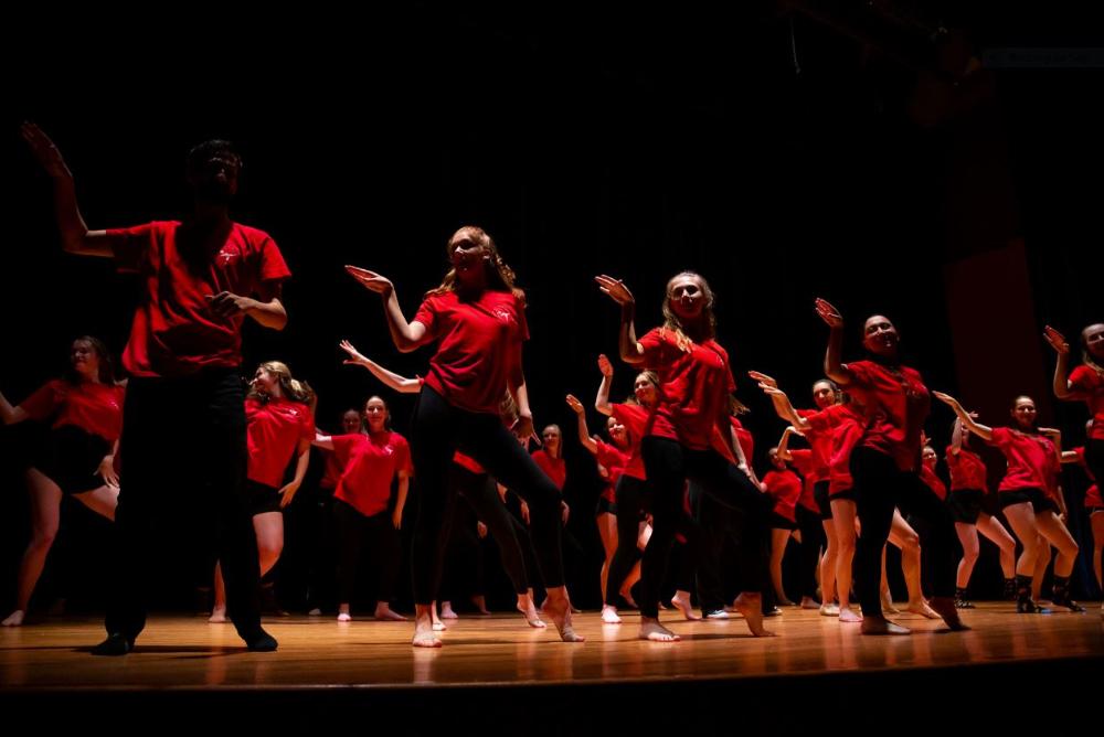 Dancers stand on stage doing a pose in unison. Dancers wear matching red shirts and are looking to their right.