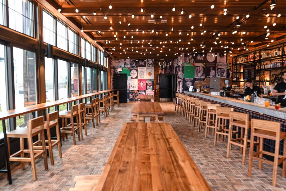 Pictured: ambient string lighting of venue. High table faces window view on the left. Bar with barstools on the right. Wooden dining tables in the middle of the venue.