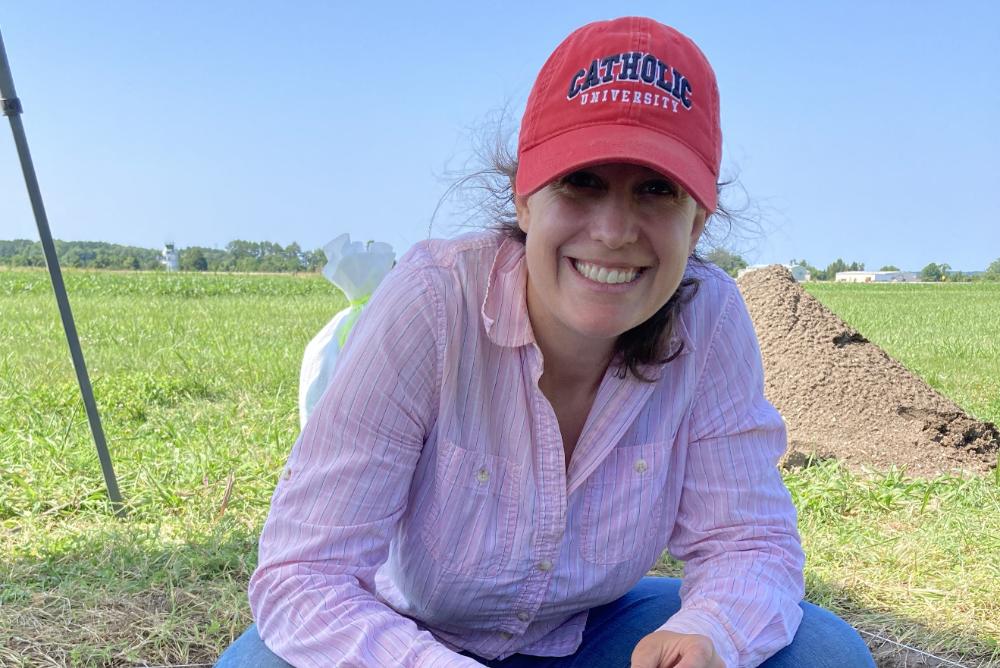 Dr. Laura Masur, crouching with an artifact, Catholic University hat, at archaeological site, blue ski, green field of grass
