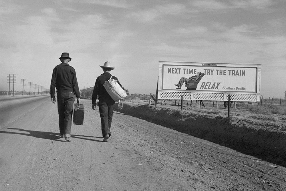 Depression-era photograph of two men walking away from the camera, carrying suitcases, next to a billboard that reads 