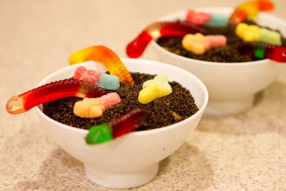 A white bowl with chocolate cake and Oreo crumbles and rainbow colored gummy worms on top.