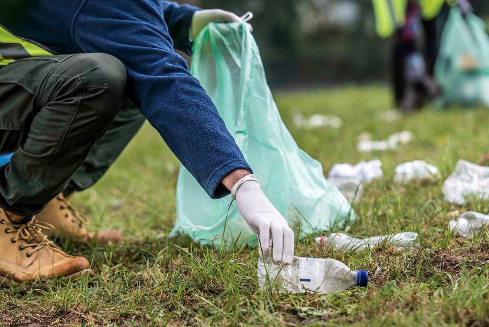 A person wearing glove and picking up trash from a grassy area. One hand is holding a green trash bag.