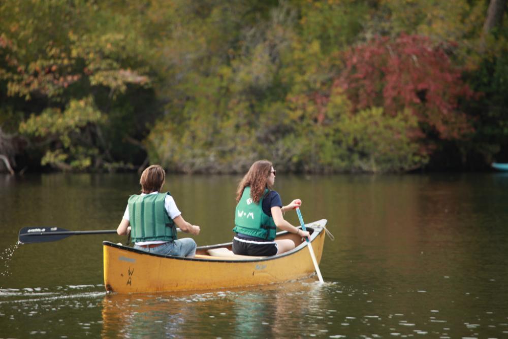 Two individuals canoeing on Lake Matoaka in a yellow canoe. They are surrounded by green and red trees.